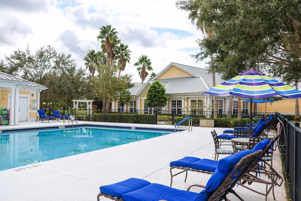 Outdoor swimming pool area with blue cushioned lounge chairs and a colorful striped umbrella. The pool is surrounded by a black metal fence, and there are palm trees and a yellow building with a metal roof in the background under a partly cloudy sky.