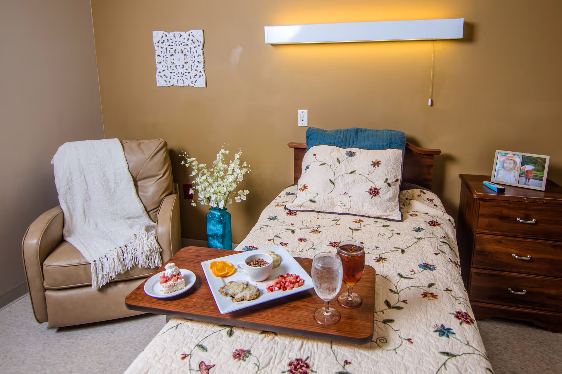 A decorated single bed in a patient room with a floral quilt, a meal tray, an armchair with a throw, and a wooden nightstand.