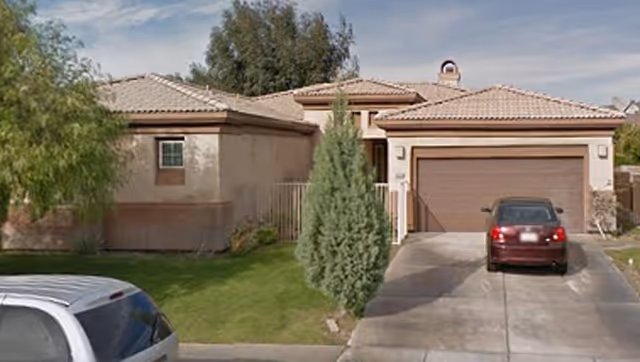 Single-story stucco house front with a two-car garage, driveway with a parked car, and a small lawn with trees.