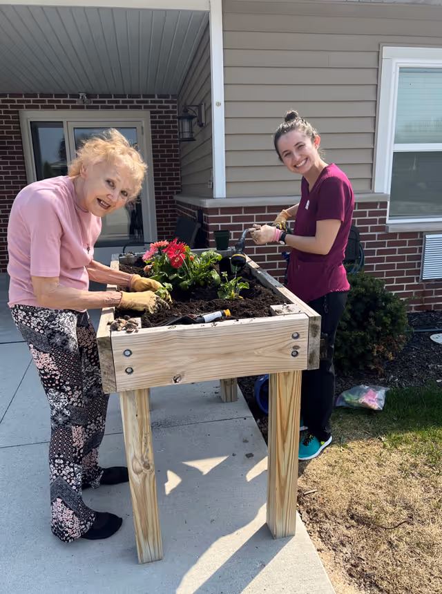 An elderly woman and a younger woman are gardening together at a raised wooden planter outside a building. The elderly woman is wearing a pink shirt and patterned pants, while the younger woman is wearing a maroon scrub top and black pants. Both are smiling and appear to be enjoying the activity. There are flowers and gardening tools on the planter, and the scene is set on a concrete patio with some grass and shrubs nearby.