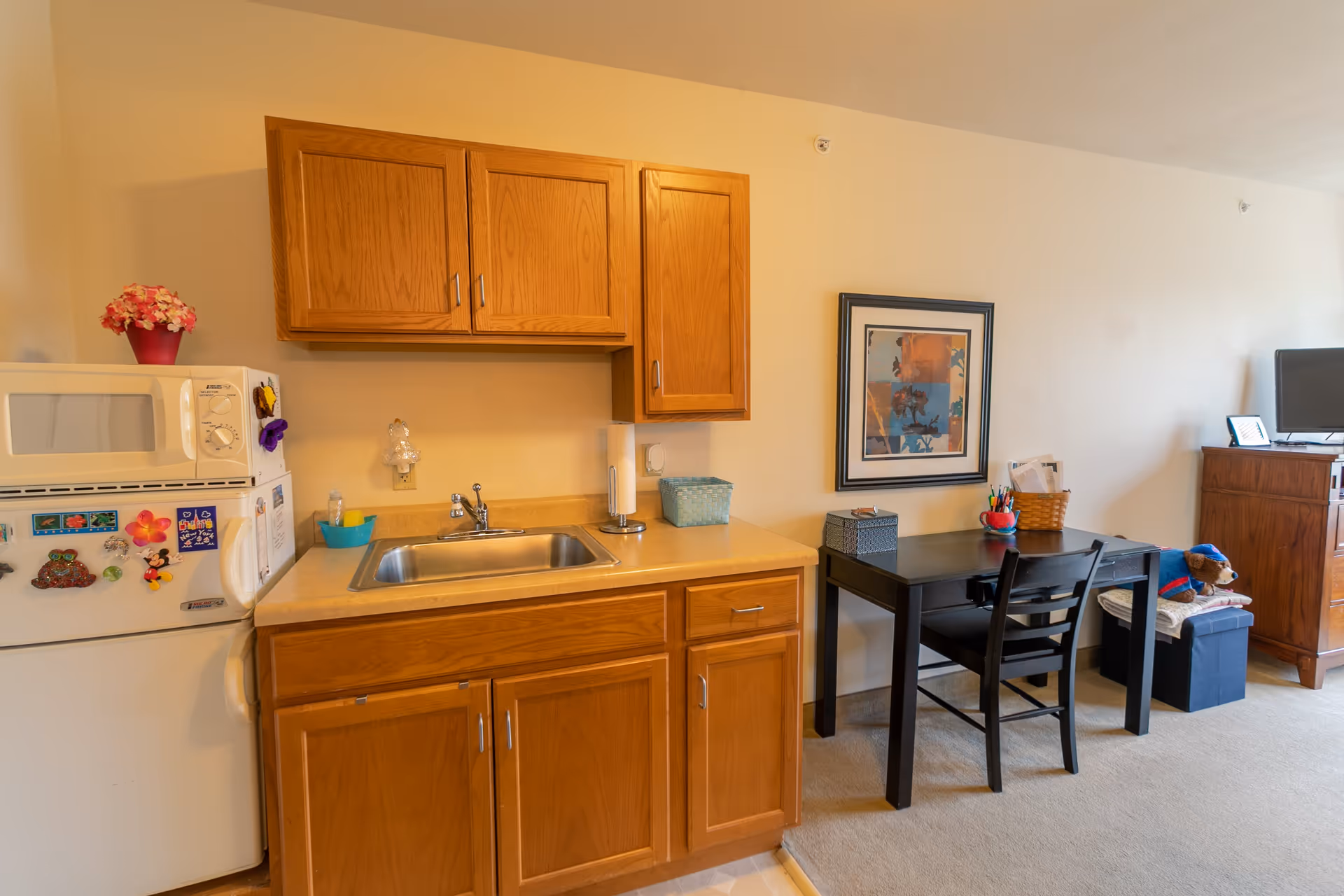 A small kitchenette area with wooden cabinets, a stainless steel sink, a microwave on top of a refrigerator decorated with magnets, and a countertop with a paper towel holder and a small basket. Next to the kitchenette is a black desk with a chair, some stationery items, a framed abstract artwork on the wall, and a TV on a wooden stand with a stuffed bear sitting on a cushion nearby.