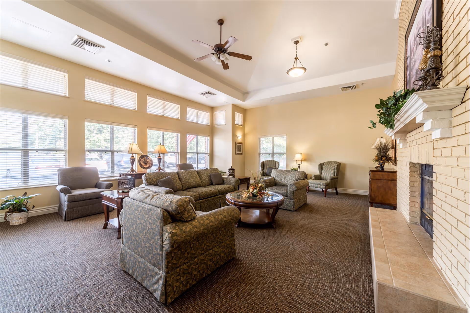 A spacious and well-lit living room with large windows along one wall. The room features multiple upholstered armchairs and sofas arranged around a round wooden coffee table with a floral centerpiece. There are side tables with lamps, a ceiling fan, and a fireplace with decorative items on the mantel. The walls are painted a soft beige color, and the carpet is dark brown.
