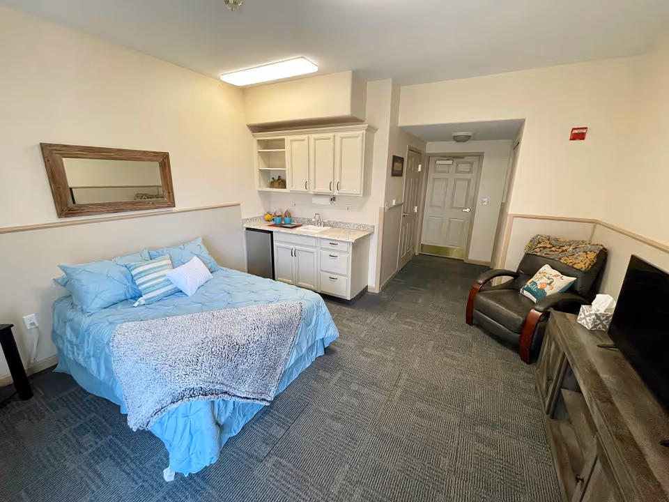 Studio-style assisted living room with a bed, small kitchenette, armchair, and TV.