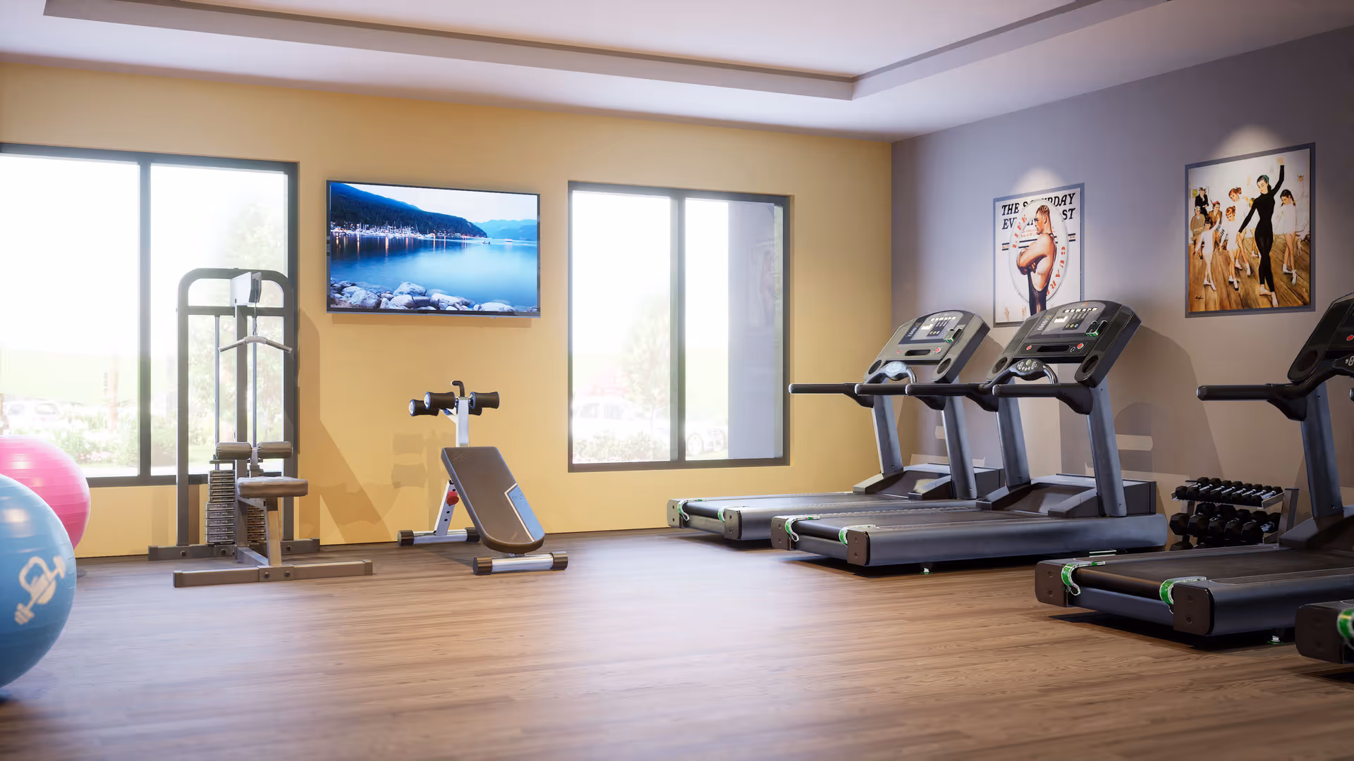 A bright fitness room with wooden flooring featuring three treadmills, a weight machine, an adjustable bench, a set of dumbbells, and two large exercise balls. The room has two large windows letting in natural light and a flat-screen TV mounted on the wall displaying a scenic lake view. There are two framed posters on the wall, one showing a ballet dancer and the other a group of dancers.