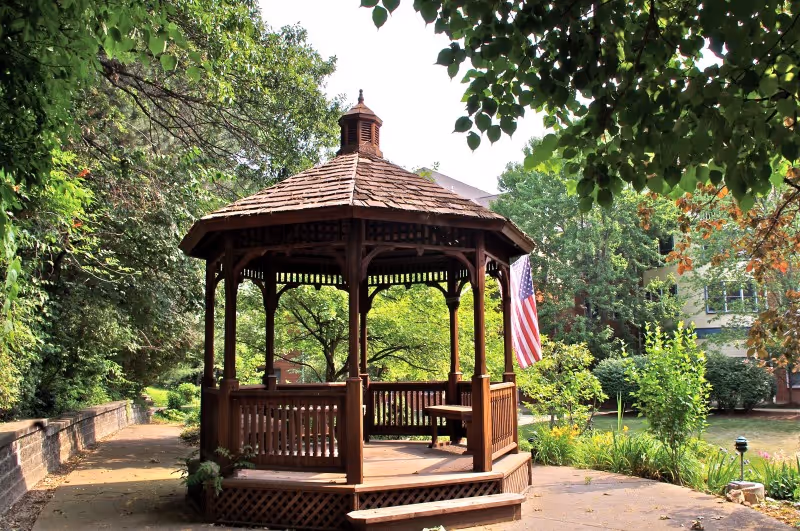 A wooden gazebo with a shingled roof situated in a garden area surrounded by trees and greenery. An American flag is hanging on the right side of the gazebo. There is a paved pathway leading to the gazebo and a building partially visible in the background.