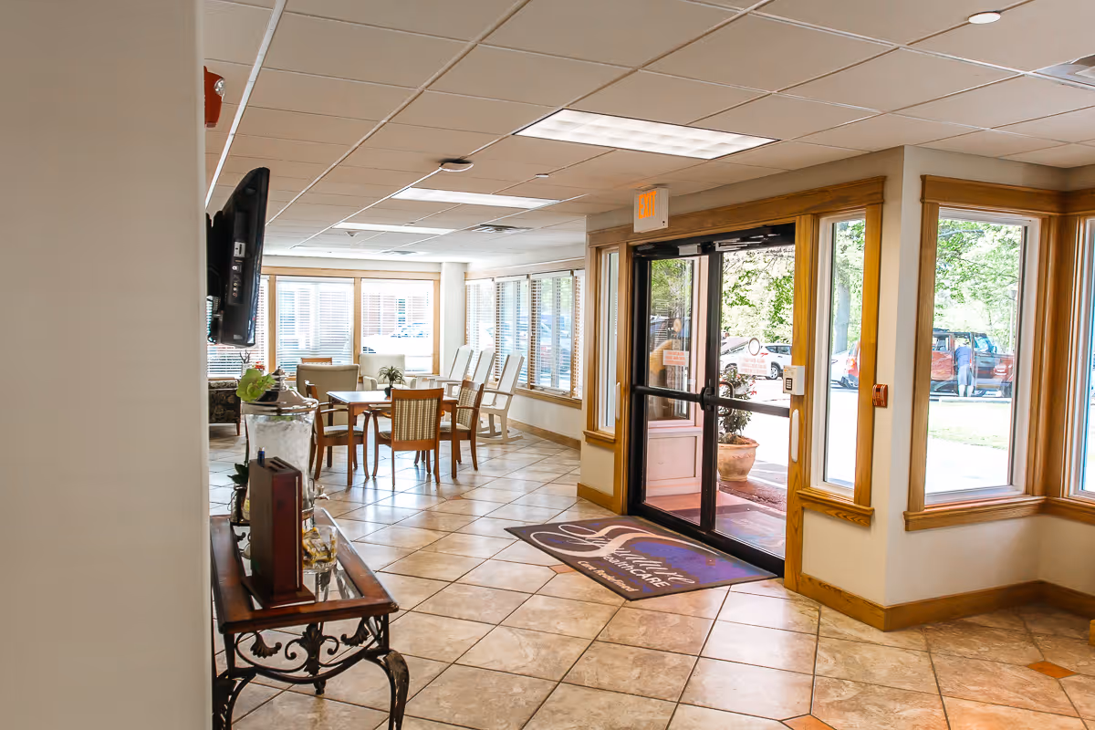Interior view of a senior living facility entrance area with large windows, a glass door leading outside, a welcome mat, tiled floor, a table with chairs, and a wall-mounted TV.
