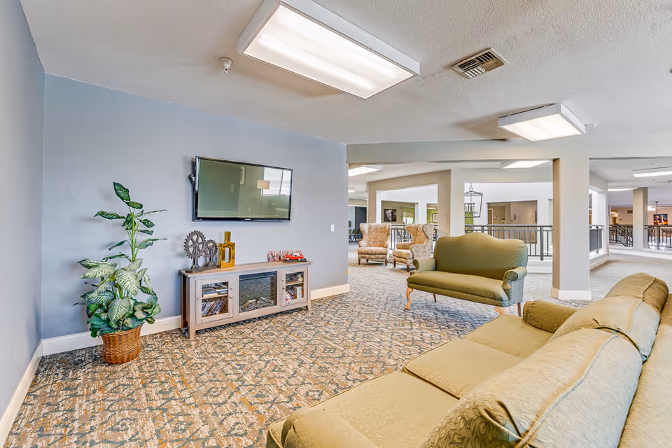 A spacious senior living common area with patterned carpet, light blue walls, and ceiling lights. The room features a mounted flat-screen TV above a wooden cabinet with decorative items and books. There is a green sofa and loveseat, a potted plant in a wicker basket, and two beige armchairs in the background near a railing overlooking another area.