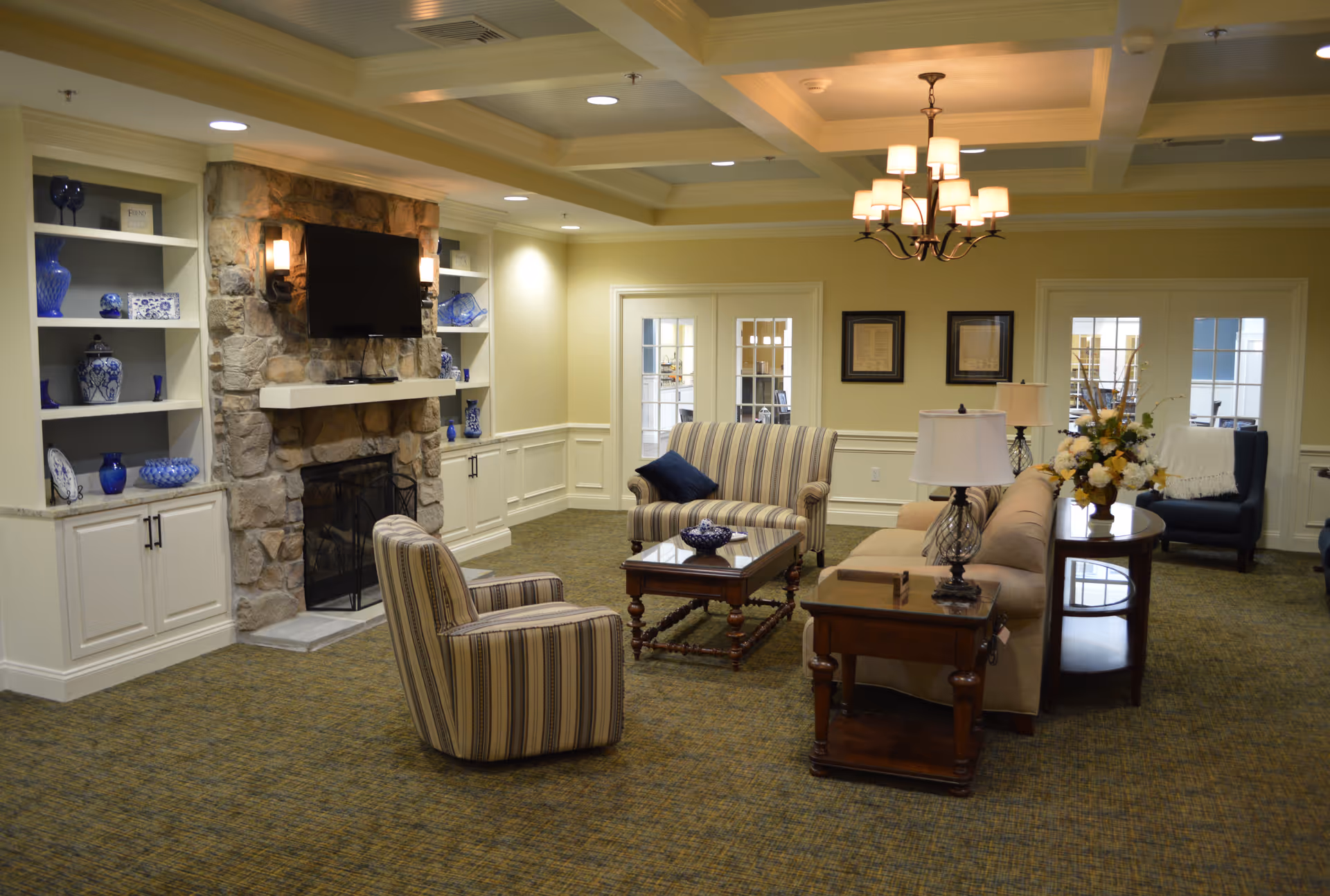 A cozy living room area in a senior living facility featuring a stone fireplace with a mounted flat-screen TV above it, built-in shelves with blue and white decorative items, a striped armchair, a striped loveseat, a beige sofa, wooden coffee and side tables with lamps, a floral arrangement, and framed pictures on the wall. The room has a coffered ceiling with recessed lighting and a chandelier, and French doors leading to another room.