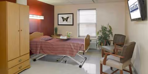 A senior care facility bedroom with a hospital-style bed, wardrobe, and chairs.