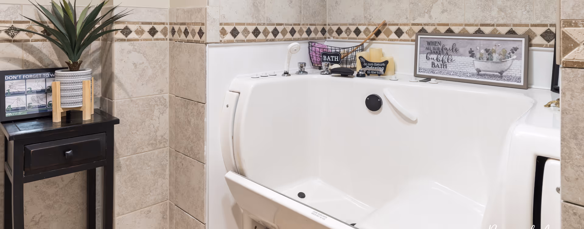 A walk-in bathtub in a bathroom with beige tiled walls and decorative border tiles. The bathtub has a built-in seat and safety handles. On the edge of the tub are bath items including soap and a small wire basket with a sign that says 'BATH'. A framed sign above the tub reads 'WHEN all else fails take a bubble BATH'. To the left of the tub is a small black wooden table with a potted plant and a sign reminding to wash hands.