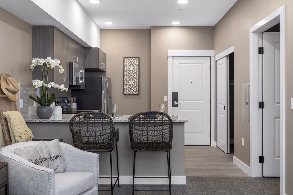 Interior view of a modern apartment kitchen and entryway. The kitchen features dark wood cabinets, a stainless steel refrigerator, microwave, and stove. A granite countertop with two black woven bar stools separates the kitchen from the living area. To the left, there is a light gray armchair with a decorative pillow and a beige throw blanket on a wooden ladder. The walls are painted beige, and there is a decorative wall hanging near the white front door.