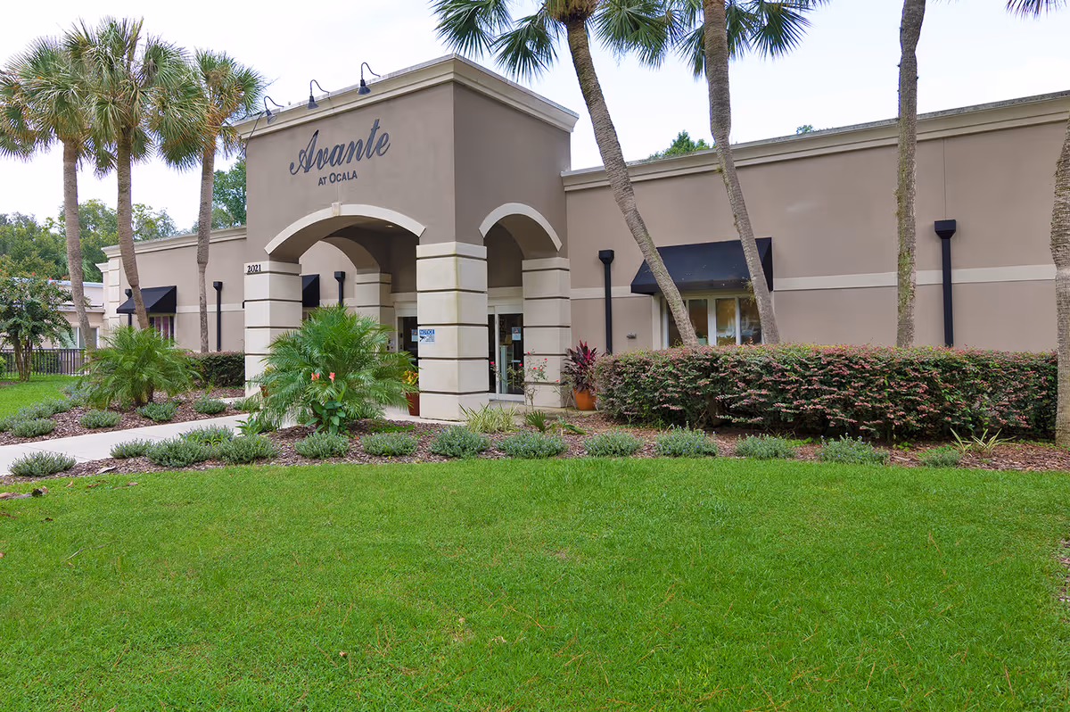 Exterior view of the Avante at Ocala senior living facility entrance with a beige building, arched entryway, palm trees, green lawn, and landscaped bushes.