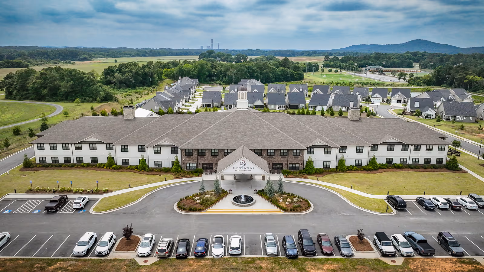 Aerial view of The Fountains in Cartersville assisted living facility showing a large two-story building with a central entrance, a circular driveway with a fountain, multiple parked cars, and surrounding greenery and smaller residential buildings in the background under a cloudy sky.