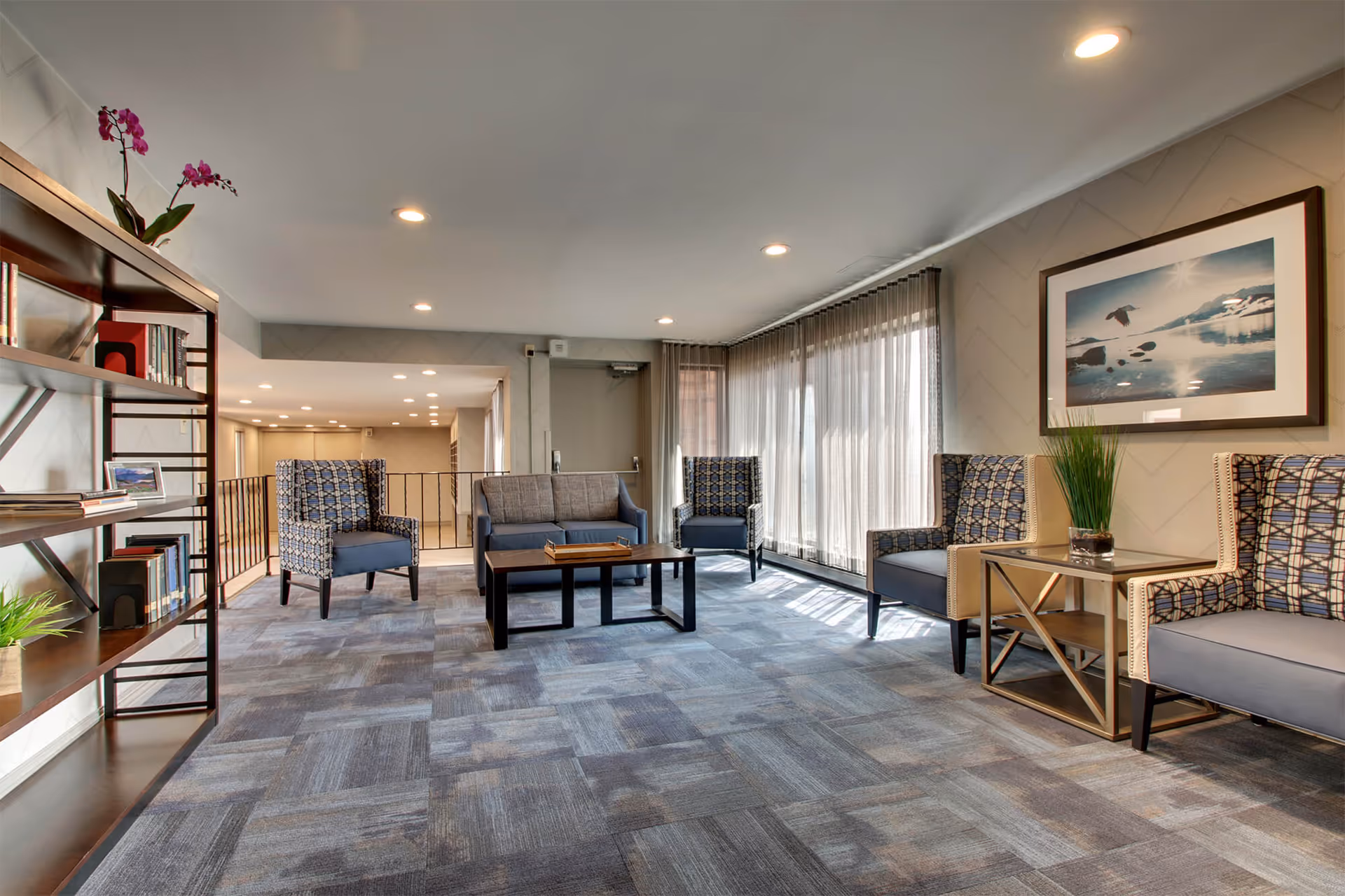 Well-lit seating lounge with armchairs, a sofa, coffee table, bookshelf, and large windows with sheer curtains.
