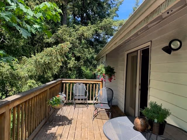 A wooden outdoor deck attached to a light-colored building with two chairs, a round table with potted plants, and hanging flower baskets. The deck overlooks lush green trees under a clear blue sky.