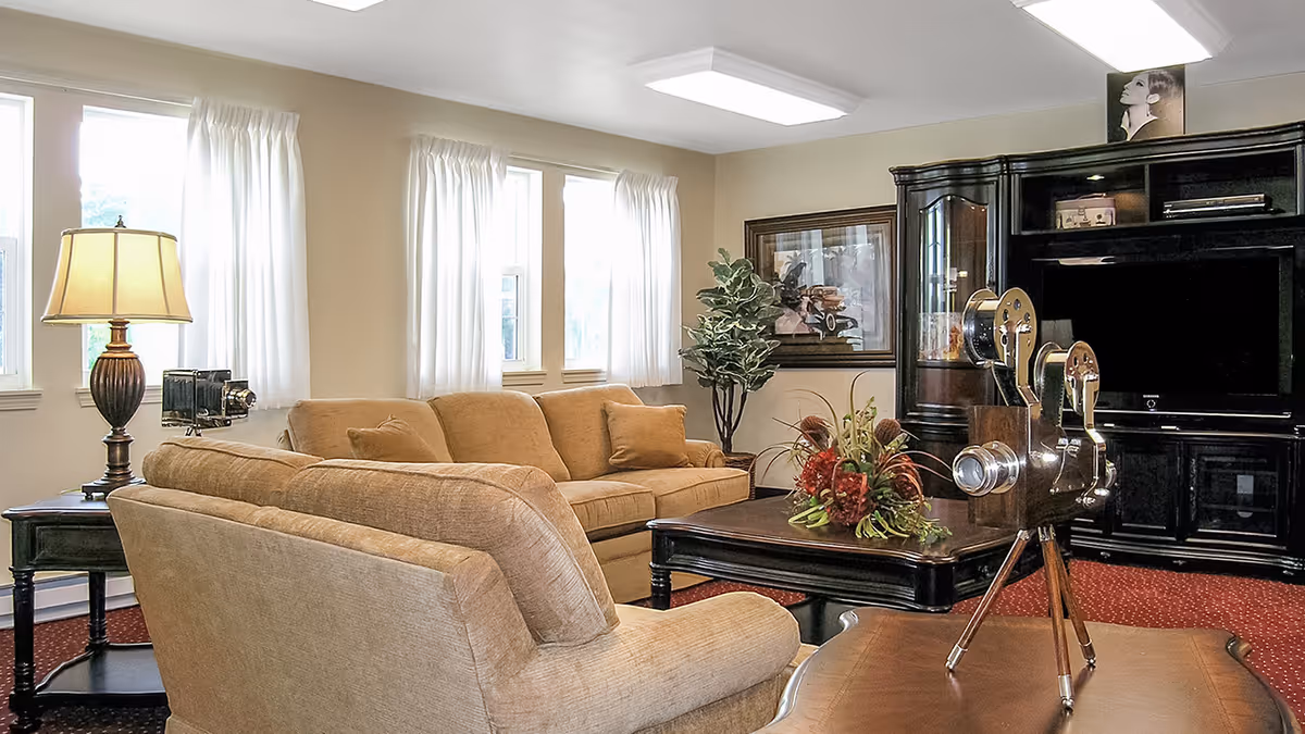 A cozy living room with beige sofas, a dark wooden coffee table with a floral arrangement, a side table with a lamp and vintage camera, and a large dark wooden entertainment center with a TV. The room has three windows with white curtains, a framed picture on the wall, and a potted plant in the corner.