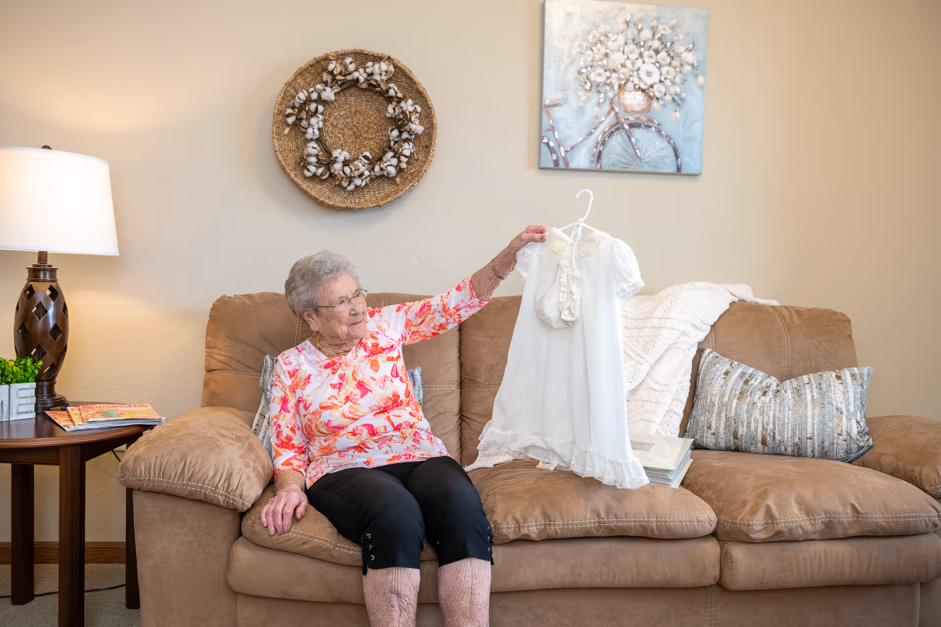 An elderly woman sitting on a brown couch in a living room, holding up a white baby dress on a hanger. The room has a side table with a lamp, some magazines, a decorative wreath on the wall, and a painting of a bicycle with flowers.