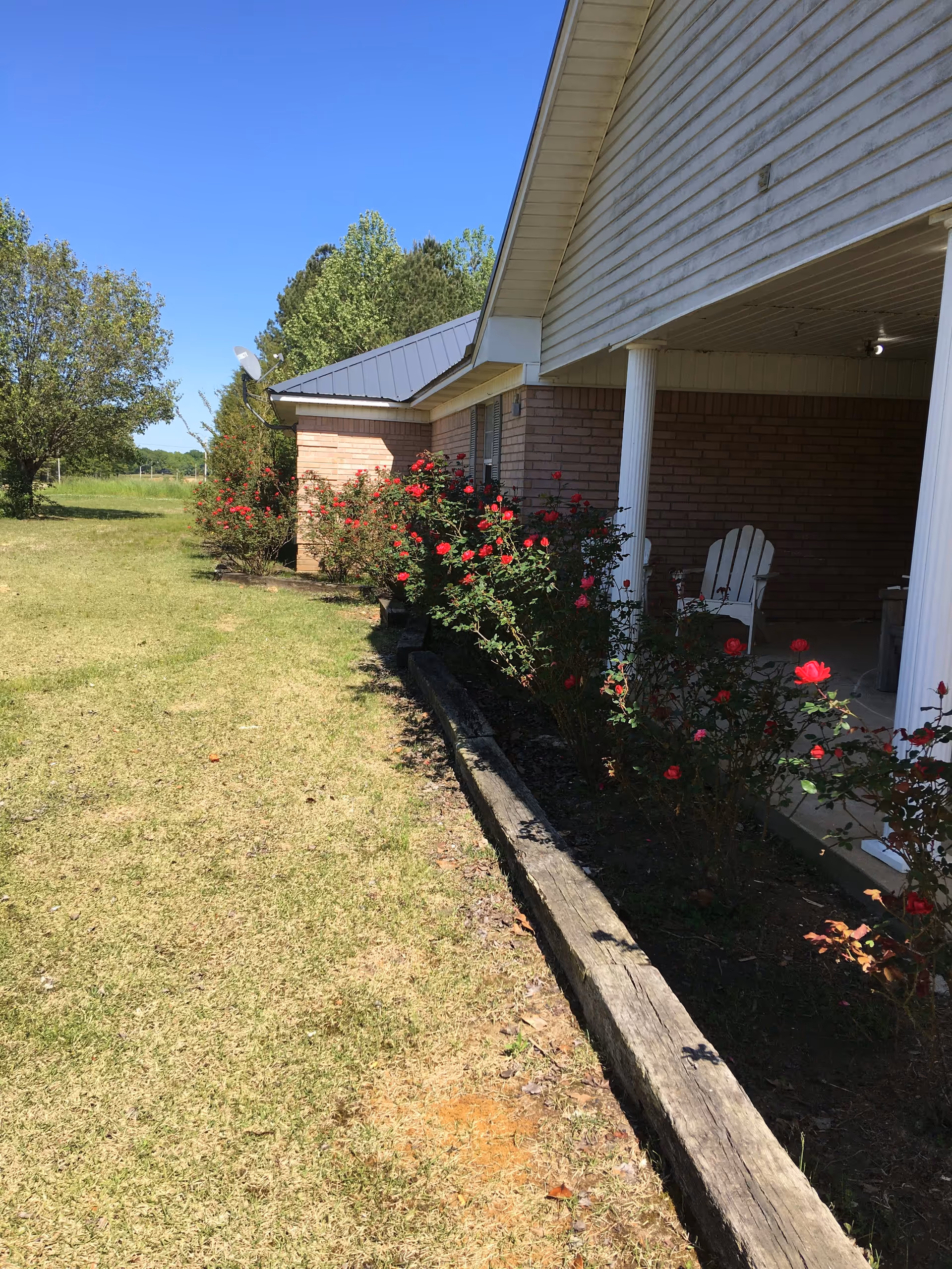 Outdoor view of a single-story brick building with a covered porch supported by white columns. There are red rose bushes planted along the edge of the porch and a grassy lawn extending outward. Trees and a clear blue sky are visible in the background.