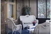 A cozy indoor seating area with white wicker furniture including a loveseat, two chairs, and a small coffee table. The loveseat and chairs have floral cushions. There is a potted plant in the corner and large windows showing trees outside.
