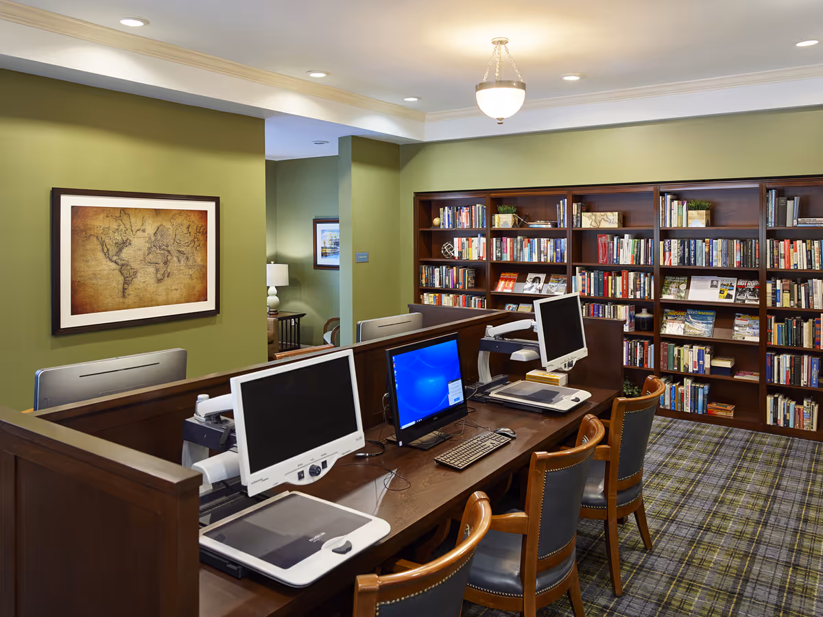 A cozy library or computer room with wooden bookshelves filled with books and magazines, a long wooden desk with three computer stations, and wooden chairs with blue cushions. The walls are painted green, and there is a framed vintage world map on one wall. The room is well-lit with ceiling lights and a hanging light fixture.
