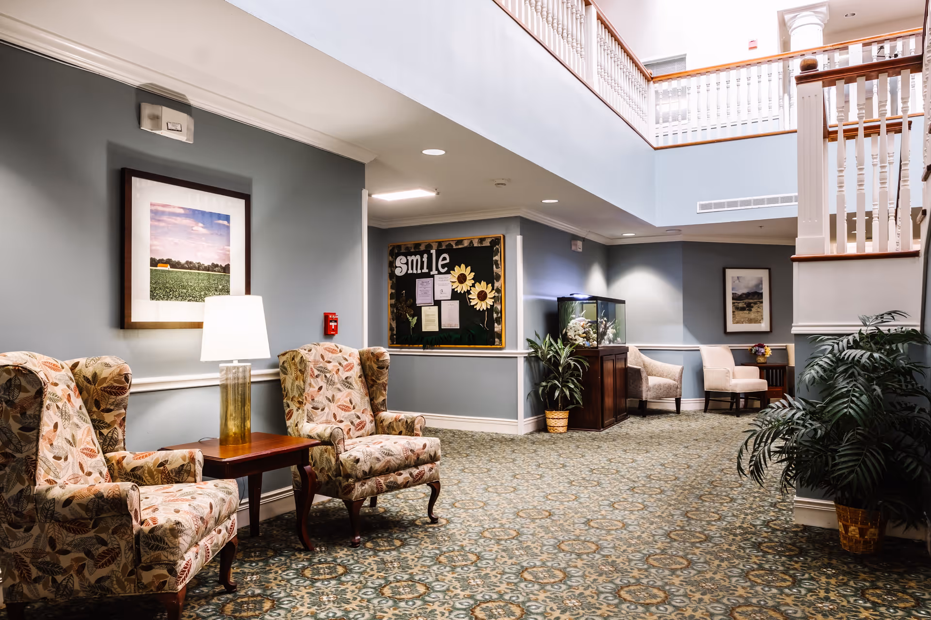 A well-lit interior common area with patterned carpet, two floral upholstered armchairs with a wooden side table and lamp between them, a bulletin board with the word 'smile' and sunflowers, an aquarium on a wooden stand, two beige chairs near a small table with flowers, framed landscape pictures on the walls, and a white railing on the upper floor.