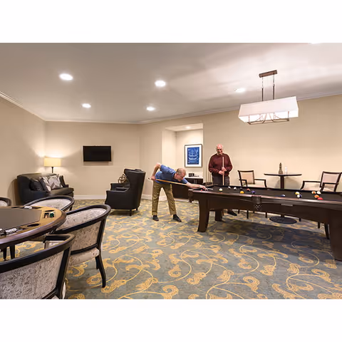 Two elderly men playing pool in a well-lit game room with a patterned carpet, several chairs and tables, a wall-mounted TV, and a lamp on a side table.