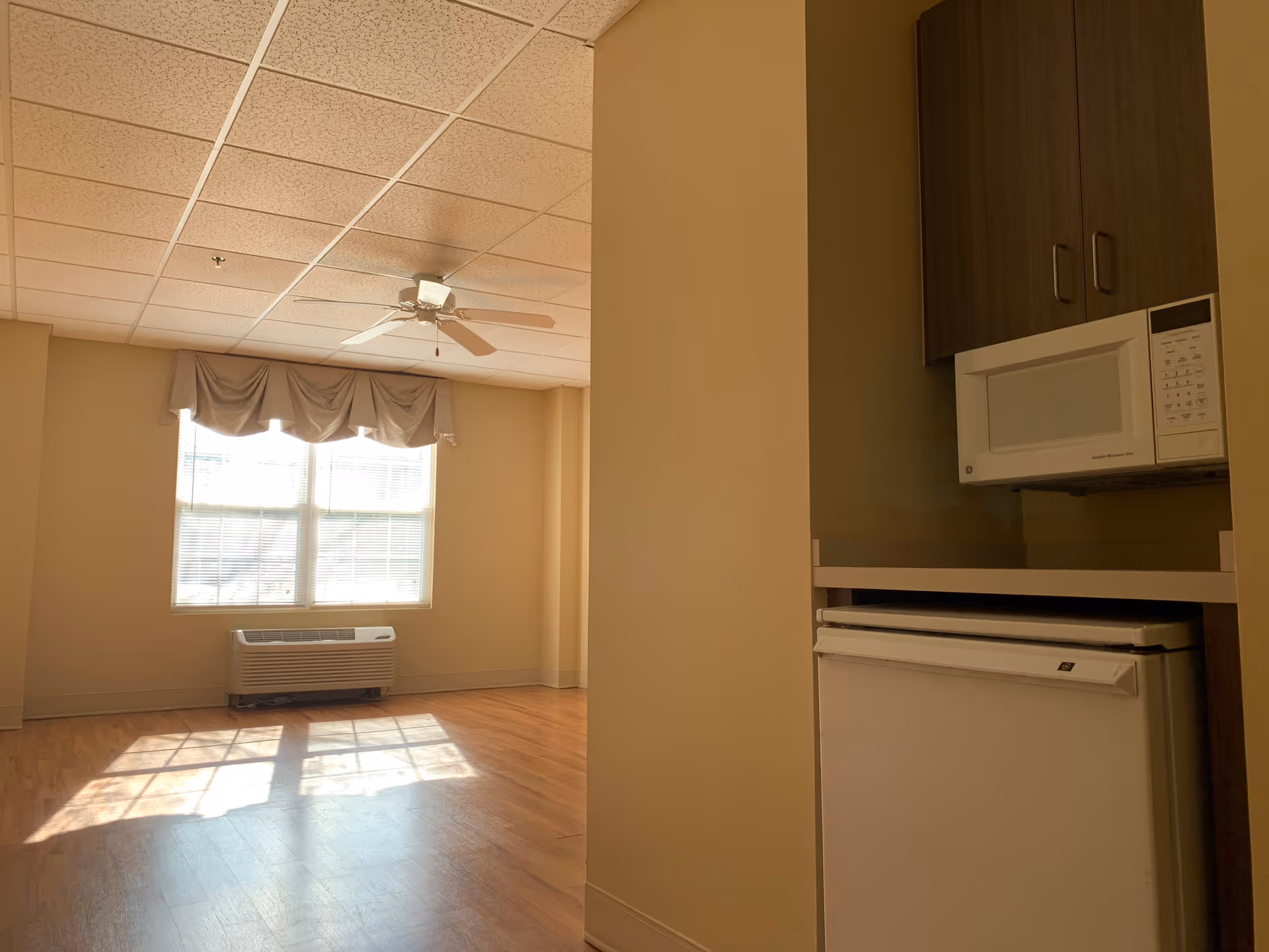 A bright room with a large window letting in sunlight, a ceiling fan, and a heating/cooling unit beneath the window. To the right, there is a small kitchenette area with a microwave above a mini refrigerator, and wooden cabinets above the microwave.