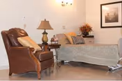 A cozy room featuring a hospital-style bed with pillows and a blanket, a wooden side table with a lamp and decorative items, a comfortable brown leather armchair with a cushion, and a framed picture on the wall.