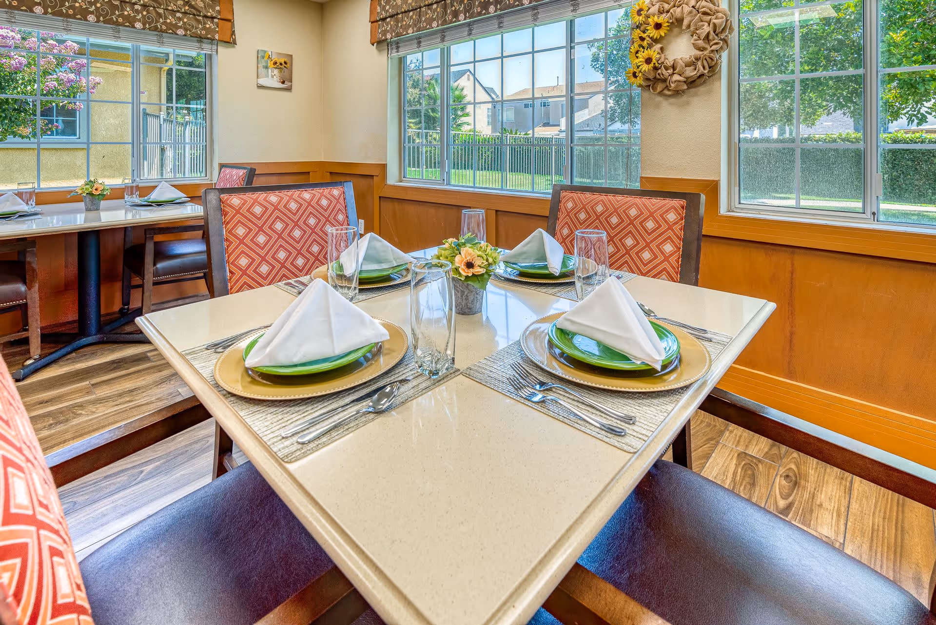 A bright dining area with a square table set for four people. Each place setting includes a gold charger plate, a green plate, a folded white napkin, a fork, knife, spoon, and a tall drinking glass. The chairs have red patterned upholstery. Large windows with floral valances let in natural light and show a view of greenery outside. A small flower arrangement is in the center of the table.