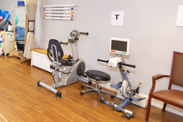 Indoor room with exercise equipment including a recumbent bike and a hand cycle machine, a rolling stool, a wooden chair, and various therapy tools on the wall and in the background. The floor is wooden and the walls are light-colored.