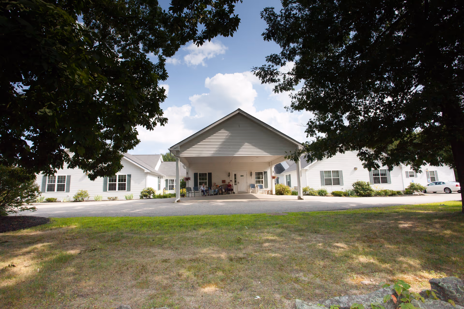 Front exterior view of a single-story white building with a covered entrance and a few people sitting on chairs under the entrance canopy. The building is surrounded by trees and grass with a partly cloudy sky in the background.