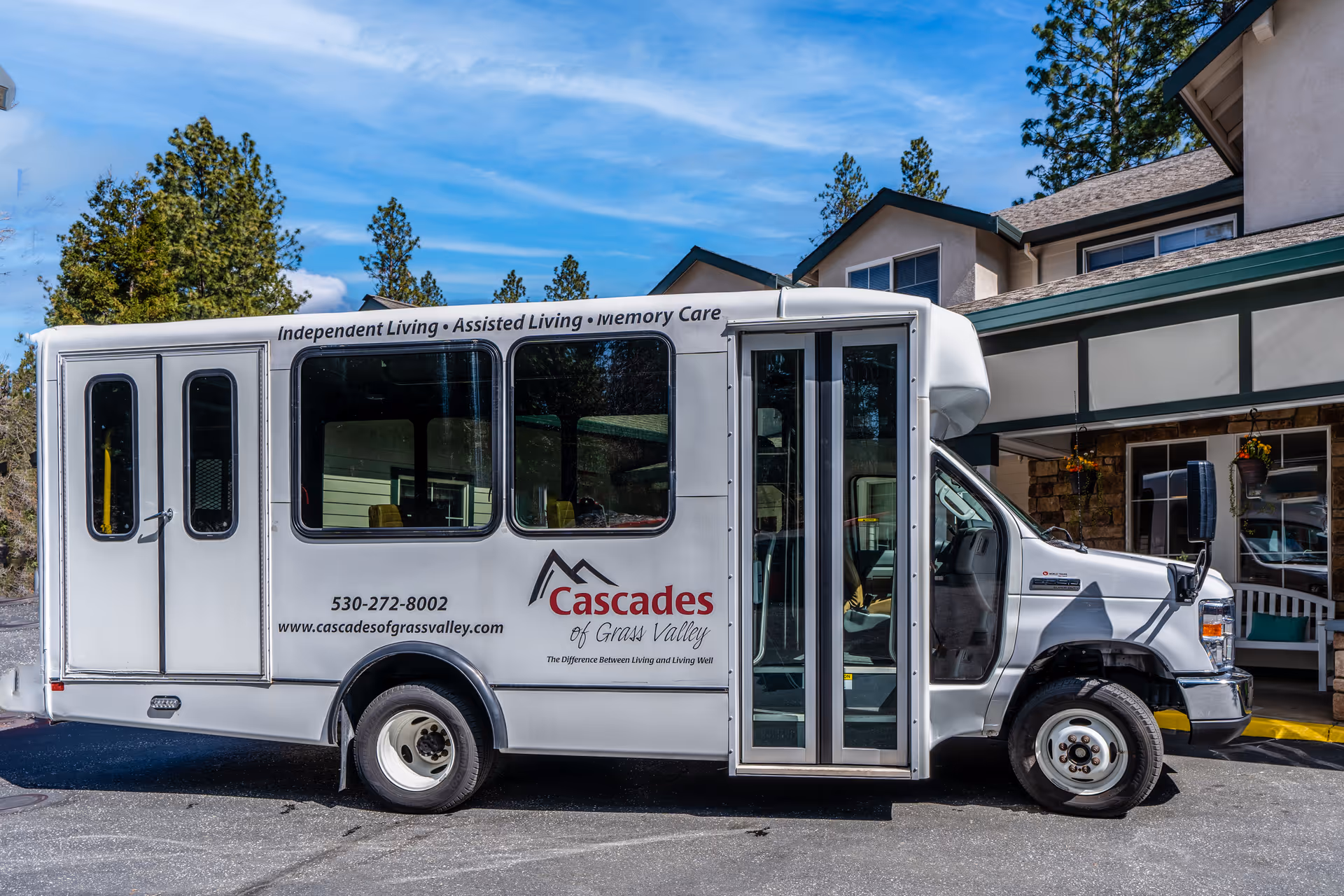 White shuttle bus parked outside a senior living facility building with signage for Cascades of Grass Valley. The bus displays services including Independent Living, Assisted Living, and Memory Care, along with contact information and the facility's website.