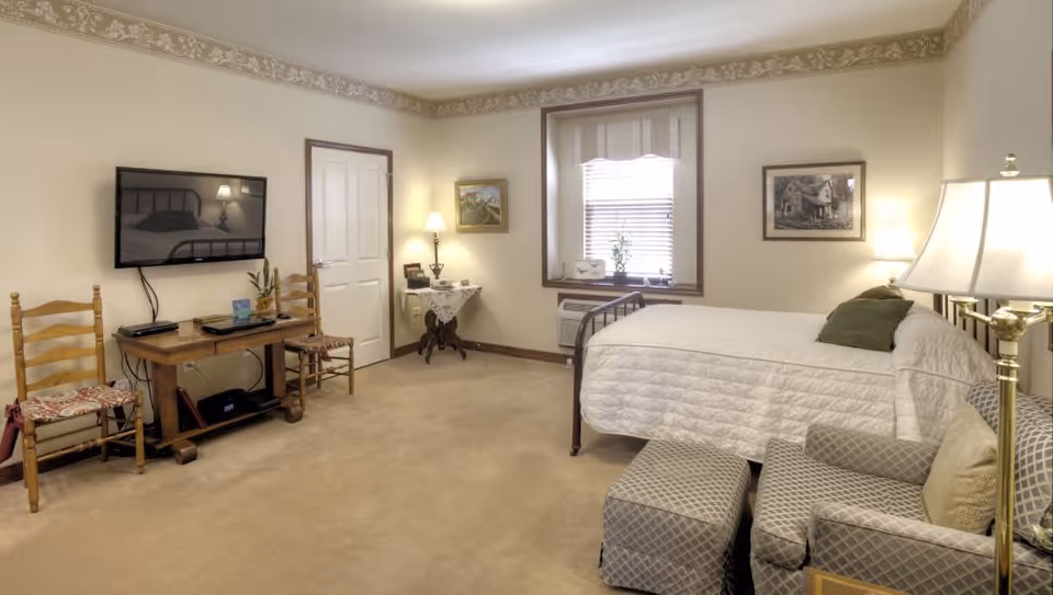 A cozy bedroom with beige walls and carpet, featuring a single bed with white bedding and a green pillow, a patterned armchair with matching ottoman, a wooden table with two chairs, a wall-mounted flat-screen TV, a window with blinds, and several lamps providing warm lighting.