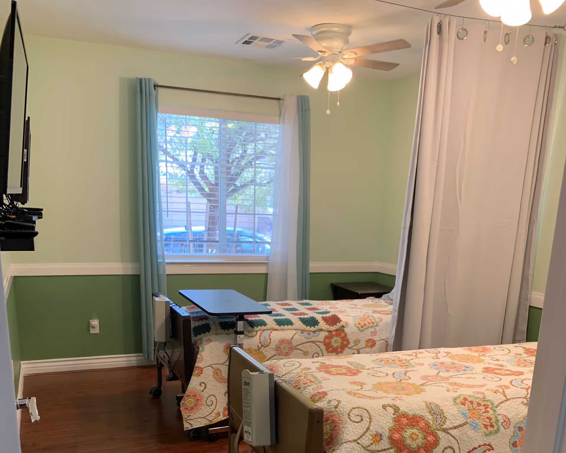 Two twin beds with floral bedspreads in a softly painted room with a window, wall-mounted TV, ceiling fan, and a privacy curtain divider.