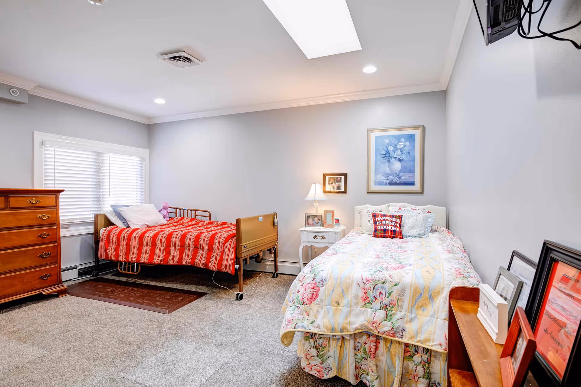 A senior living bedroom with two beds. The bed on the left has a red striped blanket and a pink stuffed animal, while the bed on the right has a floral comforter and a pillow that reads 'Happiness is being a Grandma.' There is a wooden dresser on the left, a white nightstand with a lamp and framed photos between the beds, and a small wooden shelf with framed pictures on the right. The walls are light gray with a framed floral painting above the right bed. The room is carpeted and well-lit with recessed ceiling lights and a skylight.