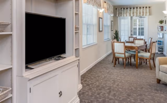 Bright communal lounge showing a TV in a white cabinet and a dining table with chairs in the background.
