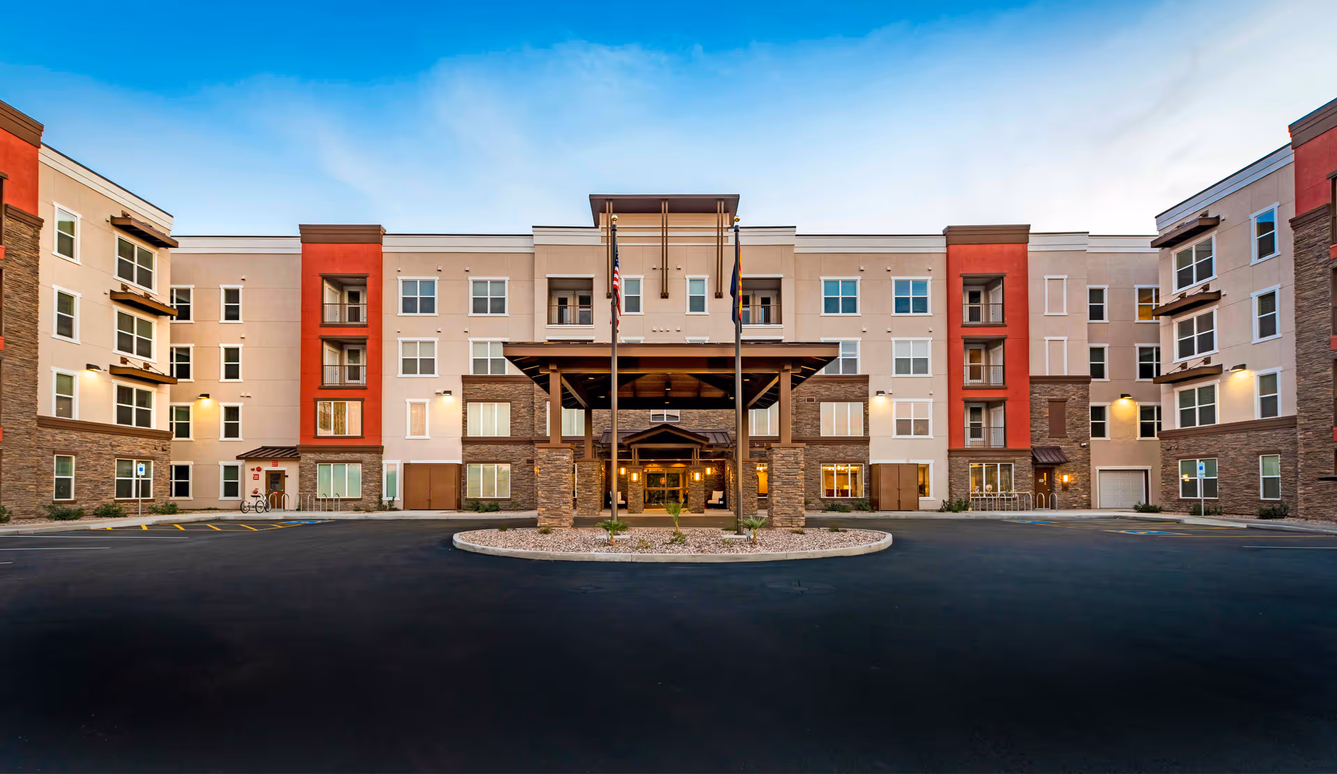 Front exterior view of Verena at Gilbert, a multi-story senior living facility with a covered entrance, stone and beige facade with red accents, and a clear blue sky above.