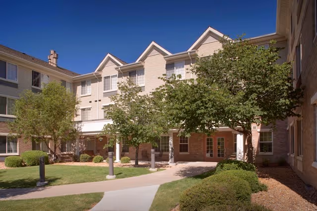 Exterior view of Clare of Assisi senior living facility showing a multi-story building with beige siding and brick accents, surrounded by green trees and bushes, with a paved walkway leading to the entrance under a clear blue sky.