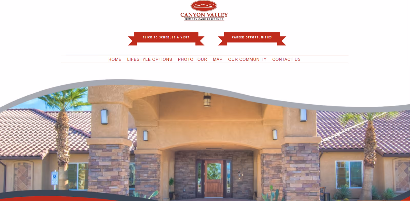 Entrance of Canyon Valley Memory Care Residence showing a covered entryway with stone pillars, a wooden door, and palm trees on either side under a clear blue sky.