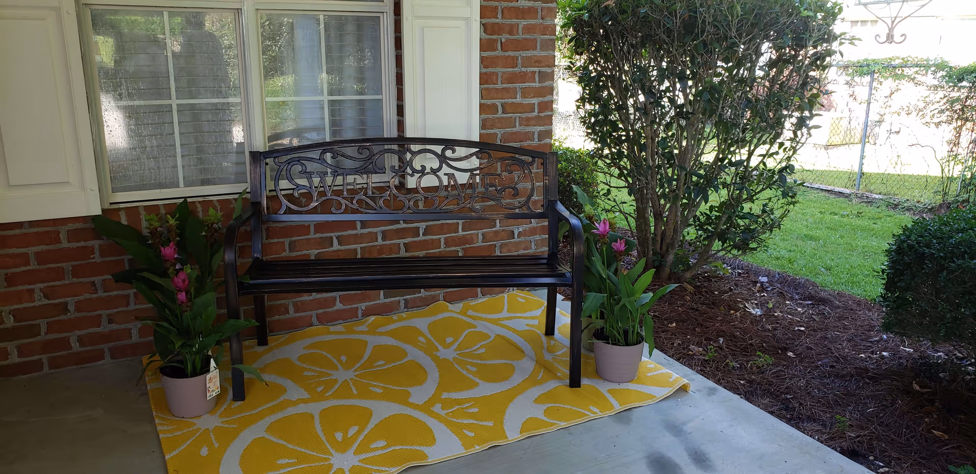 A black metal bench with the word 'WELCOME' on the backrest sits on a yellow and white patterned outdoor rug on a porch. Two potted plants with pink flowers are placed on either side of the bench. Behind the bench is a brick wall with a window and white shutters. To the right, there is a garden area with bushes and a chain-link fence.