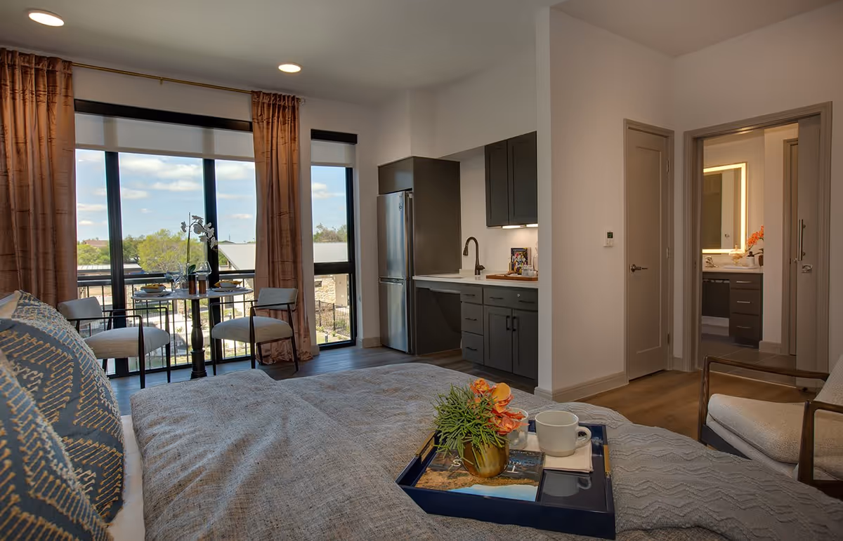 Interior view of a senior living facility room at The Hacienda at Georgetown featuring a bed with decorative pillows and a tray with a plant and a cup. The room has large windows with brown curtains, a small dining table with two chairs near the window, a kitchenette with dark cabinets and a stainless steel refrigerator, and a doorway leading to a bathroom with a lit mirror.