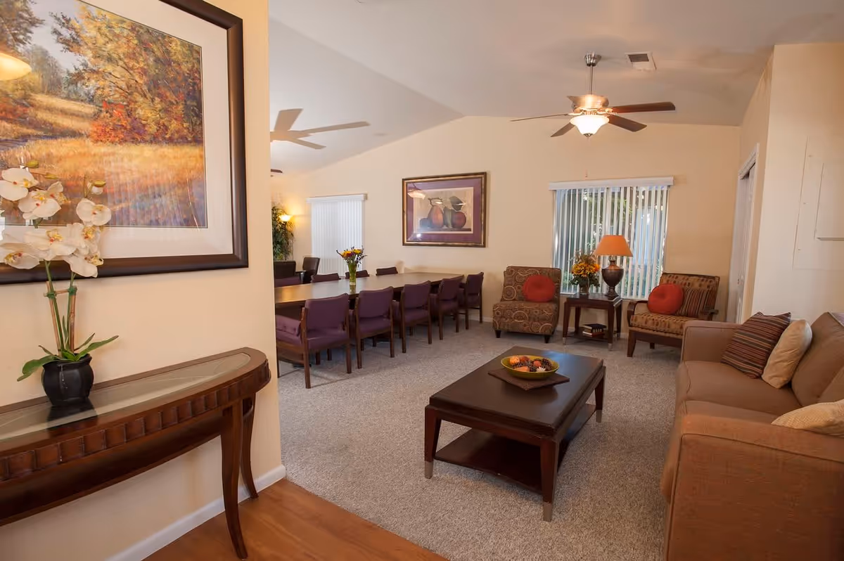 Spacious communal living room and dining area with sofas, armchairs, a coffee table, and a long dining table under ceiling fans.