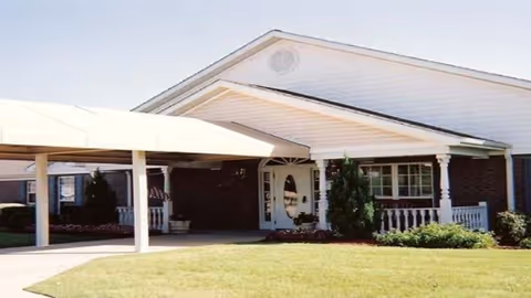 Front entrance of a single-story brick senior living facility with a covered drive-up canopy, white columns, and a small lawn.