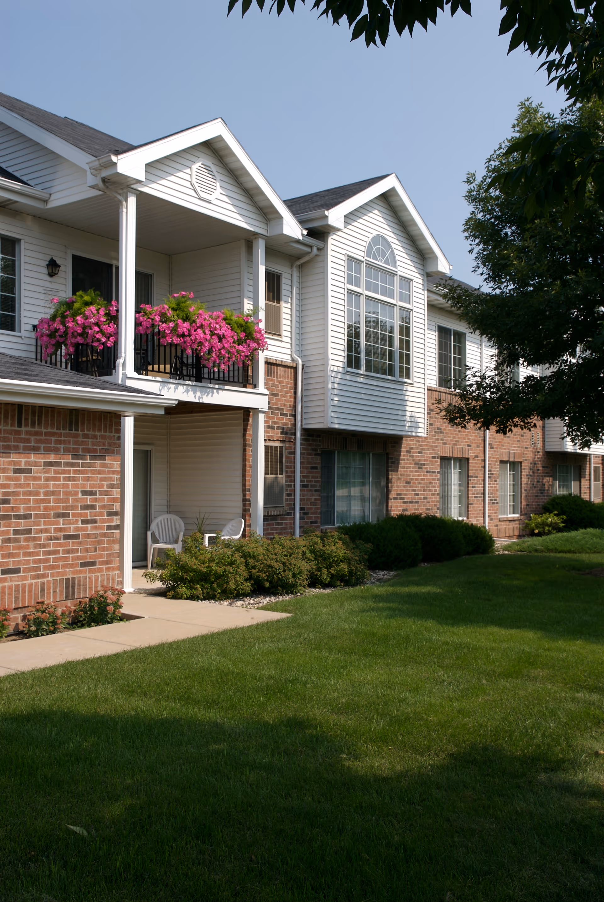 Two-story brick and siding apartment building exterior with balconies, pink flowers, and a manicured green lawn.