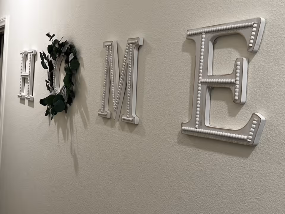 A wall decorated with large white letters spelling the word 'HOME'. The letter 'O' is replaced by a green leafy wreath. The wall is painted a light beige color.
