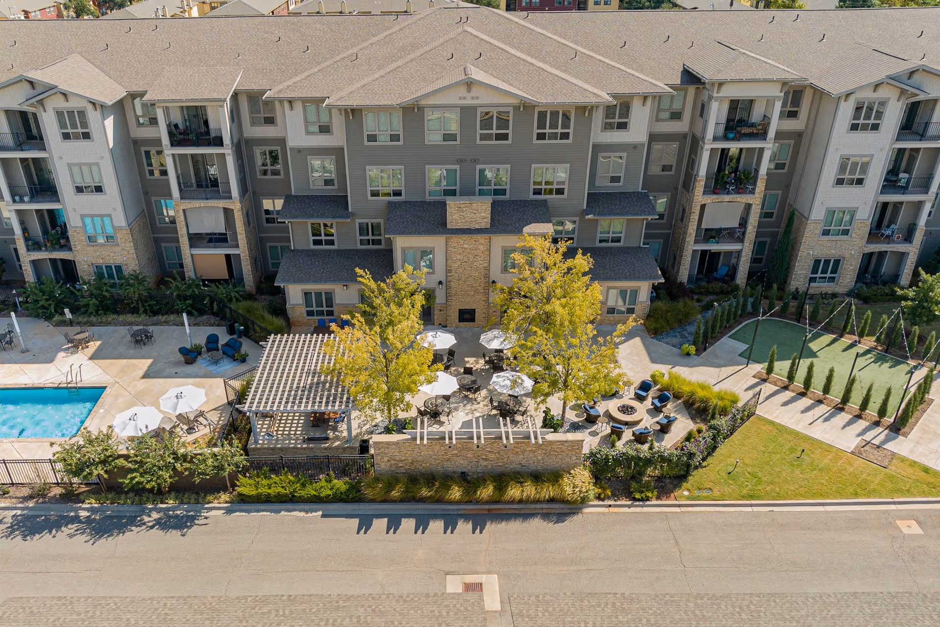 Aerial view of Album Quail Springs senior living facility showing a multi-story residential building with balconies. In front of the building is a landscaped outdoor area featuring patio tables with umbrellas, seating around a fire pit, a pergola, a swimming pool with lounge chairs, and a bocce ball court. Trees and shrubs are planted throughout the outdoor space.