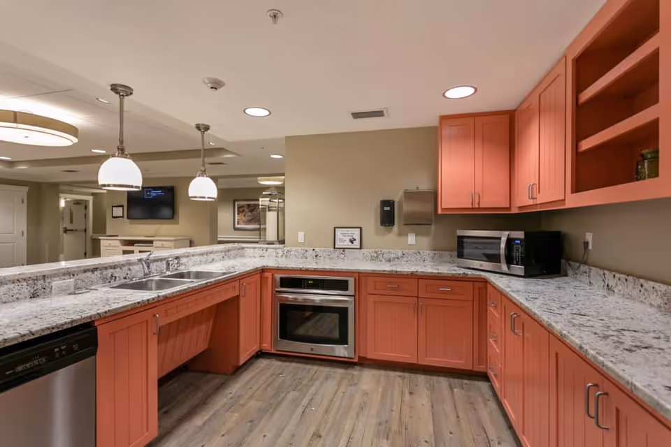 A modern kitchen area with coral-colored cabinets and drawers, white and gray granite countertops, a built-in oven, a microwave, a double sink, and stainless steel dishwasher. The floor is wood-style laminate, and there are two pendant lights hanging from the ceiling. The background shows a hallway with a TV mounted on the wall and a framed picture.