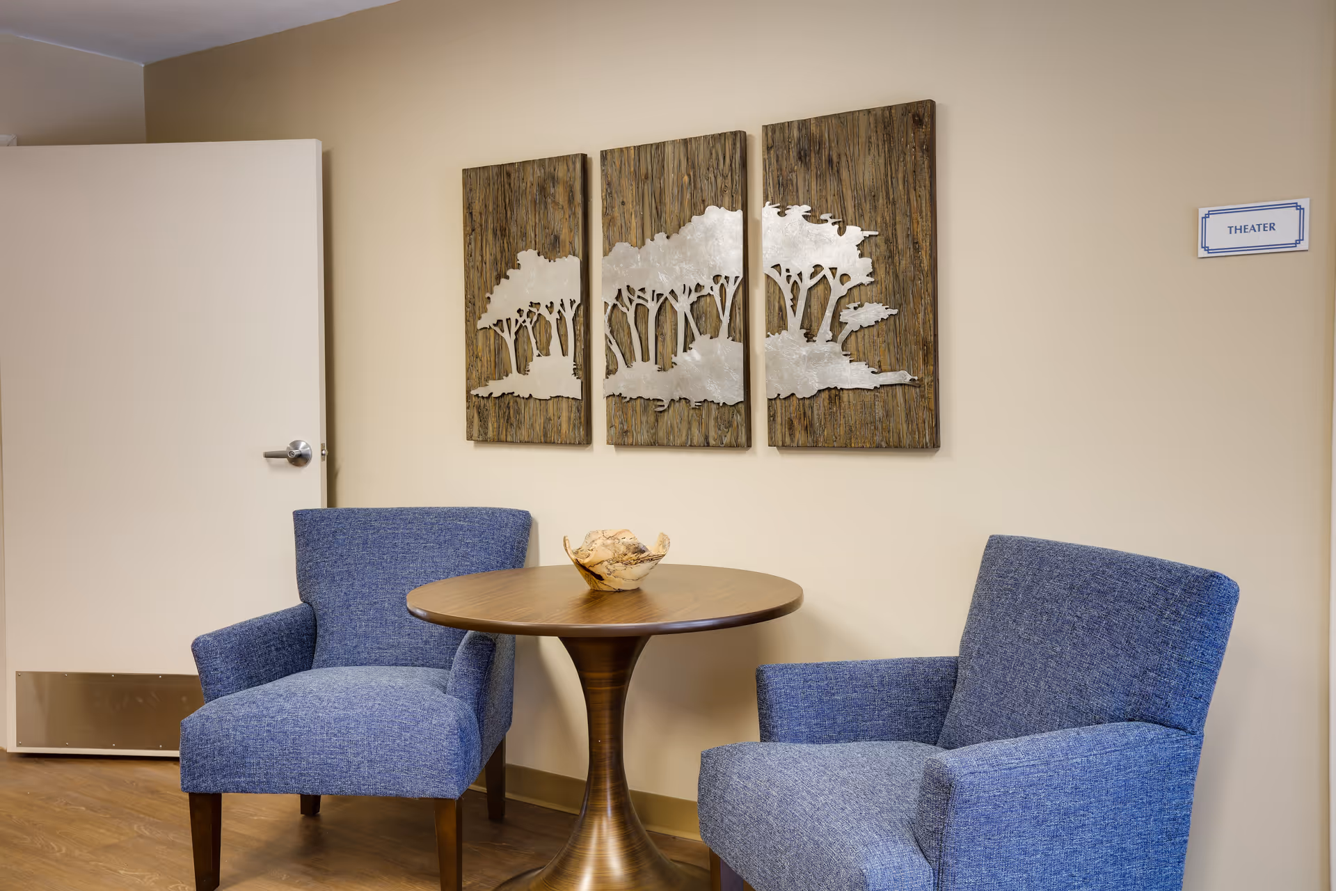 A small seating area with two blue upholstered armchairs and a round wooden table between them. On the table is a decorative bowl. Behind the seating area, there is a three-panel wall art piece depicting trees, and a door is partially open to the left. A small sign on the wall to the right reads 'THEATER'.