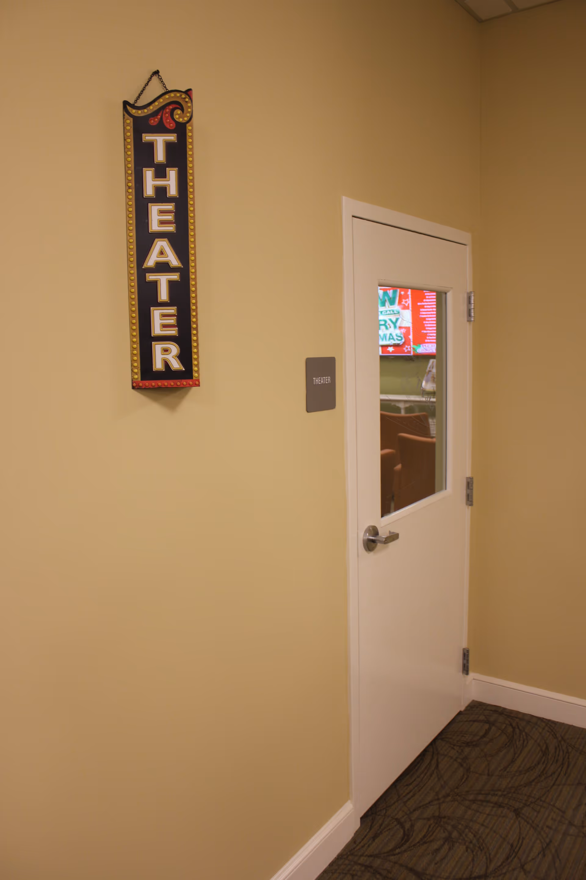 A beige hallway wall with a vertical decorative sign that reads 'THEATER' hanging next to a white door with a window. Through the window, a TV screen and some chairs are visible inside the theater room.