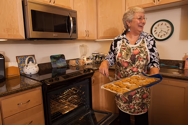 An elderly woman wearing glasses and a floral apron is smiling while holding a tray of freshly baked cookies in a kitchen. The kitchen has wooden cabinets, a microwave above the stove, and various kitchen items on the counter including a blender and a teapot. A decorative plate with bird illustrations is mounted on the wall.